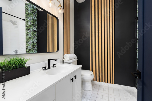 A bathroom with a white cabinet and countertop, blue door, black a wall with wood slats, and a leaf pattern wallpaper.