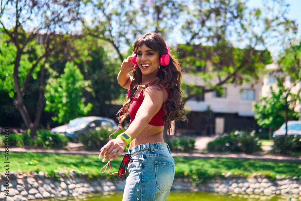 Naklejka premium A beautiful Latina woman dances with rhythm and agility. She wears pink headphones, makeup and jeans. She is dancing in an outdoor park in a retro style. She exudes attitude, style, freedom and beauty