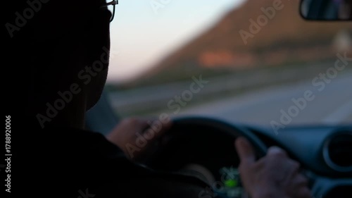 Senior man driving a car on a winding road. Elderly man wearing glasses silhouetted against sunset light, driving along winding road with approaching car, capturing late evening journey