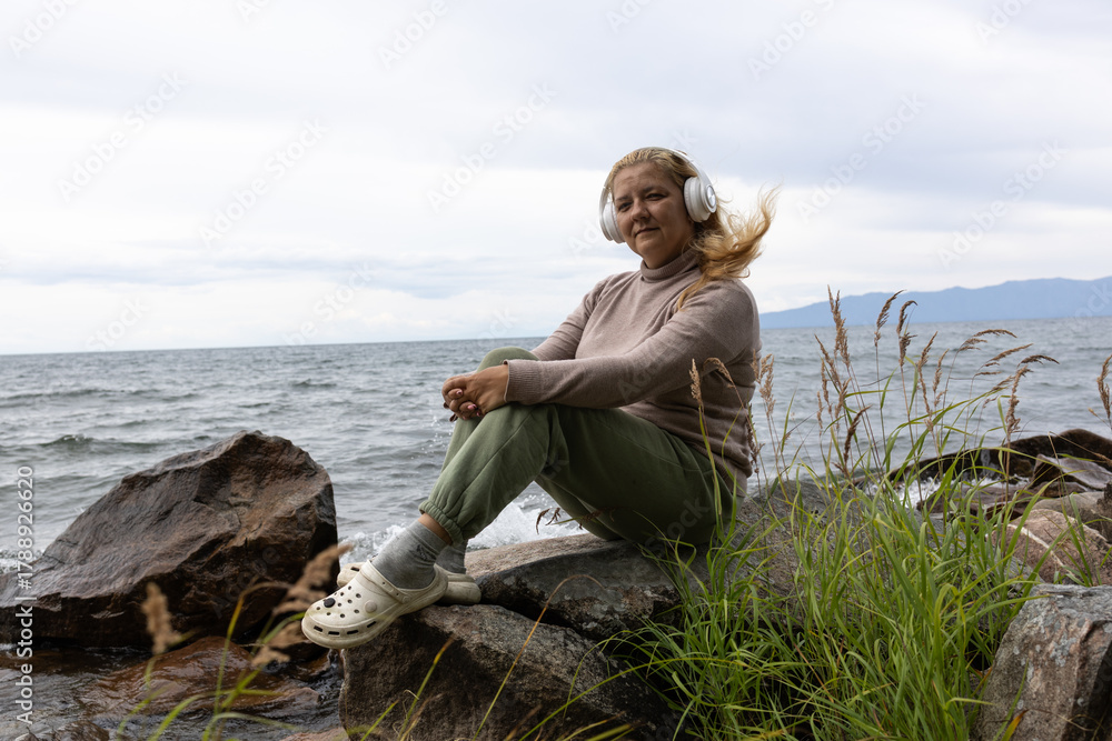 Fototapeta premium A white woman with long blonde hair listens to music with headphones, relaxing on the shore of Lake Baikal.