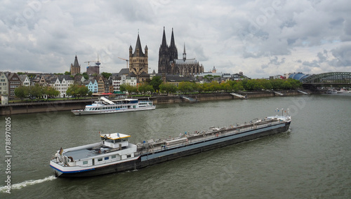 View of transport ship on the Rhine River heading North going past Old Town Köln (Cologne) waterfront, St. Martin Church, and,The Kölner Dom as seen from the Deutzer Brüke.