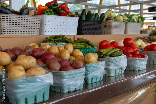 A display at a farmer's market of fresh, raw vegetables.There are multiple baskets with white and red potatoes, tomatoes, green beans, kohlrabi, onions, cauliflower, and red, green and orange peppers.