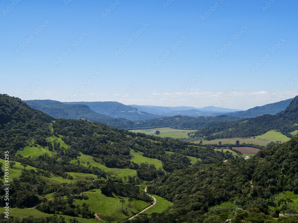 Fototapeta premium Panoramic view of green mountain valley with forested hills and clear blue sky in the countryside of southern Brazil, ideal for nature and travel themes