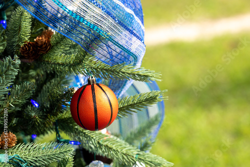 A small glass basketball Christmas ornament hanging from an evergreen tree. There's a blue striped ribbon with glitter. The tree has cones growing on the branches. The string lights are bright blue.