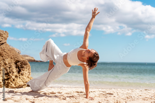 Woman in white activewear balances in Side Plank yoga pose on sunlit beach, reaching top arm toward sky. She enjoys outdoor exercise, combining strength and stability by the sea.