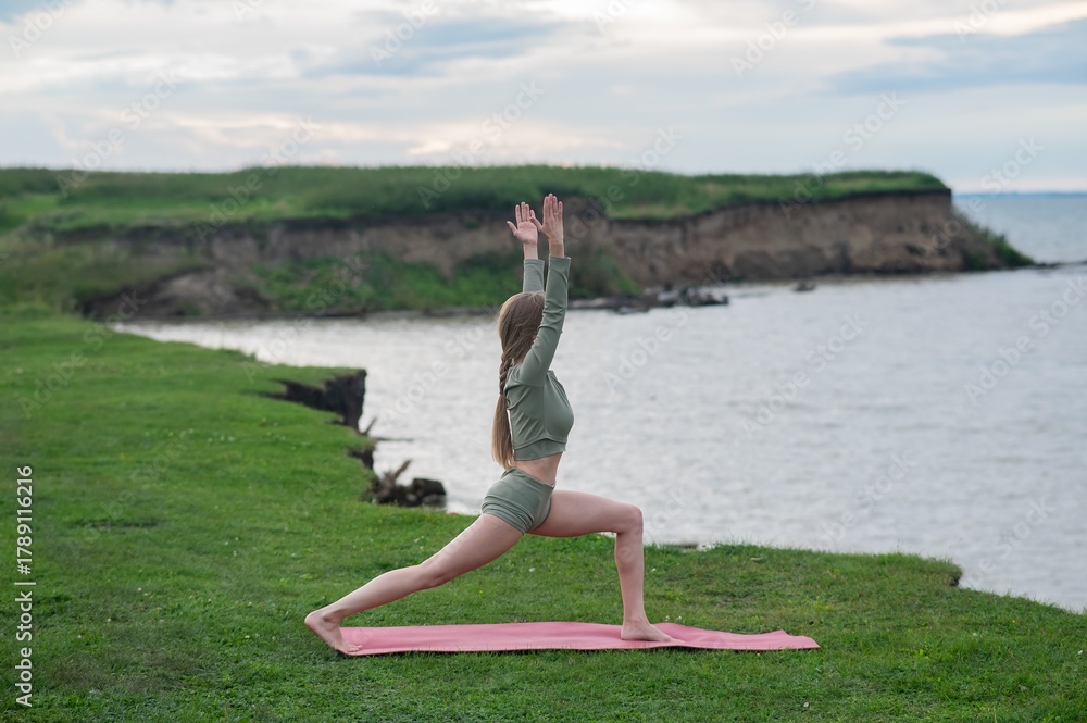 Fototapeta premium Caucasian woman doing yoga on the river bank. Warrior pose.