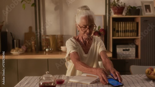 Medium shot of cheerful elderly Caucasian woman with gray hair, living with chronic diabetes ailment in old age, placing smartphone to sensor patch, checking blood glucose and writing in notebook