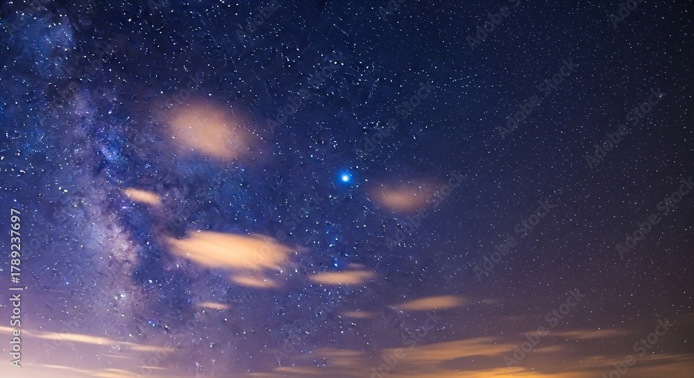 Obraz premium Astrophotography of a stunning night sky, featuring the faint glow of the Milky Way, scattered stars, and a single, intensely bright blue star. Wispy clouds are subtly lit by a warm glow from below th