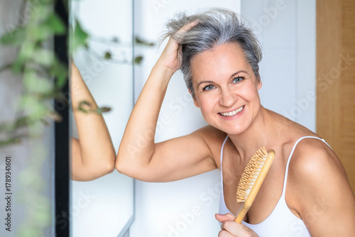 European beautiful and smiling middle-aged woman combing her gray hair near the mirror