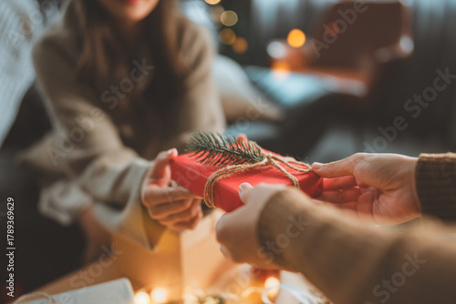 Close up of people exchanging Christmas gifts, hands giving and receiving present box wrapped in red paper with pine decoration, festive holiday season celebration with warm light and cozy home.