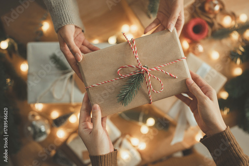 Hands exchanging Christmas gift box wrapped in brown paper with pine decoration and red white string, festive holiday season celebration with warm lights, friendship and happiness in home, New year