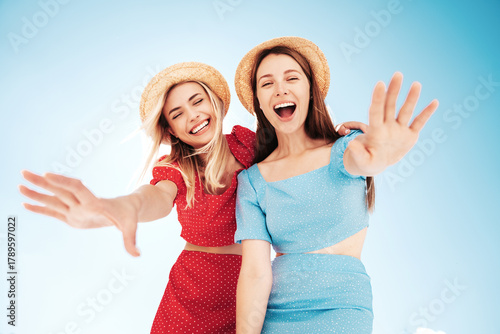 Two young beautiful smiling hipster woman in trendy summer same clothes. Carefree women in street in hats. Positive models at sunset. Cheerful and happy. Bottom view. They look at camera from above