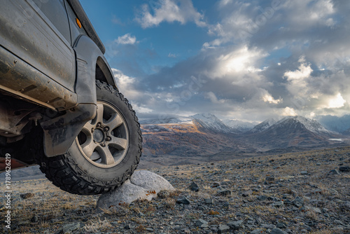 An off-road vehicle with a muddy tire posed on a rock during the golden hour. Extreme close-up on the aggressive tread with snow-capped mountains in the background. Adventure and power.
