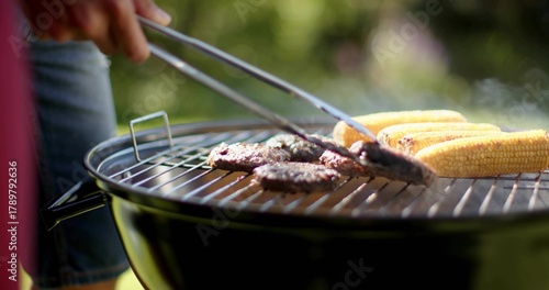 Grilling burger patties and corn on charcoal kettle grill in backyard, wearing denim and red top