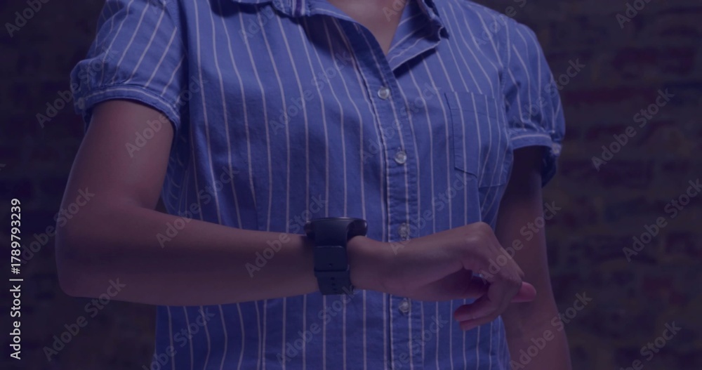 Naklejka premium Pointing, woman adjusting smartwatch on left wrist at brick wall in blue shirt under blue light