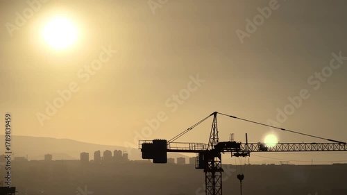 Construction crane against the backdrop of a city with a bright sun . A crane is in the sky above a city at day The crane is tall and is in the middle of the sky