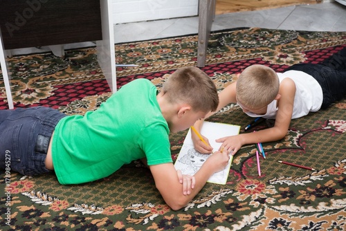 Brothers share a creative moment coloring a picture together, lying on an ornate rug beneath a table. Bonding time fosters imagination.