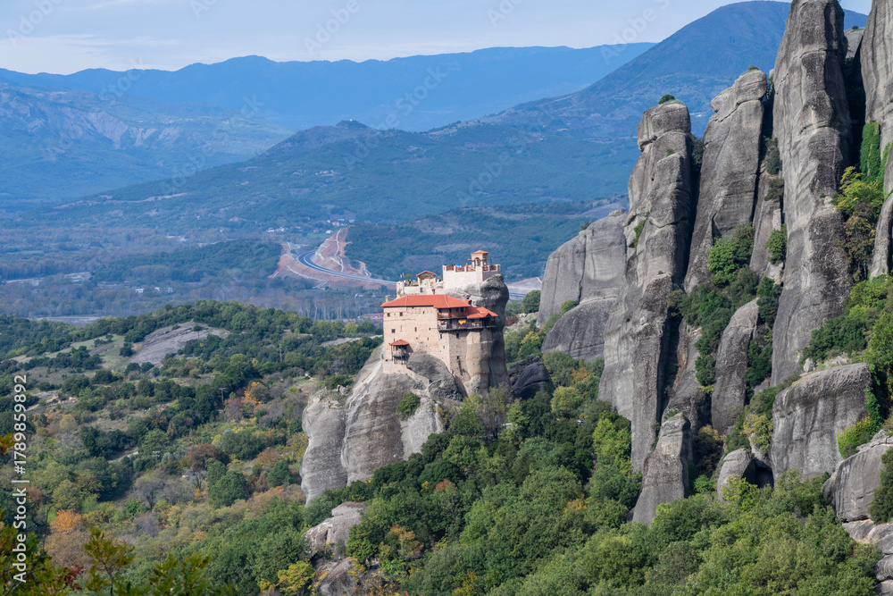 Fototapeta premium A monastery with red-tiled roofs crowns a steep rock pillar in Meteora, surrounded by lush forests and dramatic cliffs.