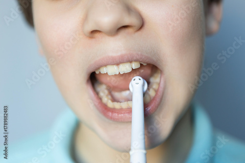 Child brushes teeth with a toothbrush featuring a smiley face while wearing a blue shirt in a well-lit room