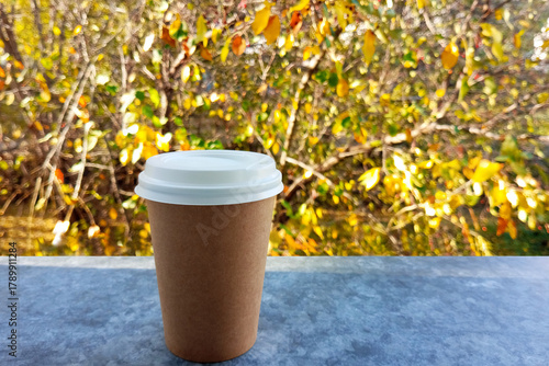 Brown craft glass with white lid with coffee on grey tabletop against autumn foliage background in sunlight.