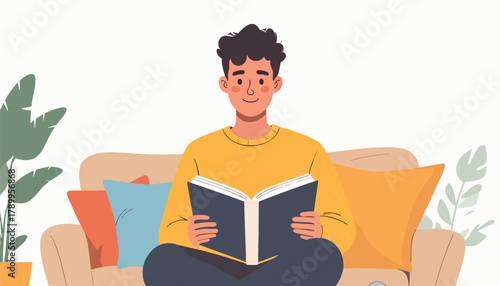 A young man sits on a couch, cross-legged, reading a book with a smile.