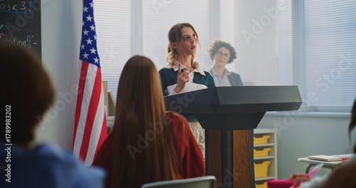 American Female Student Delivers Speech With US Flag in Front of Classmates Sitting Desks. Concept Civic Education, Public Speaking Skills, and Student Voice in American Education System. Dolly Shot.