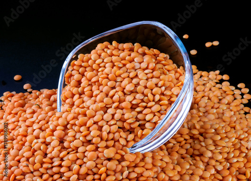 Red lentils spilling out of a glass cup on a black background. Side view. Lots of lentils around the cup.
