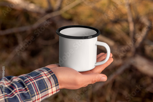 Mockup of a white enamel mug with a black rim in the hand of a man in a flannel shirt, on a background of trees, outdoor.