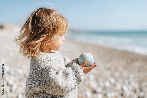 Child holding globe on sandy beach looking at the ocean waves