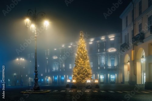 Canvas-taulu A brightly lit Christmas tree stands in a foggy, empty city square in Asti, Italy