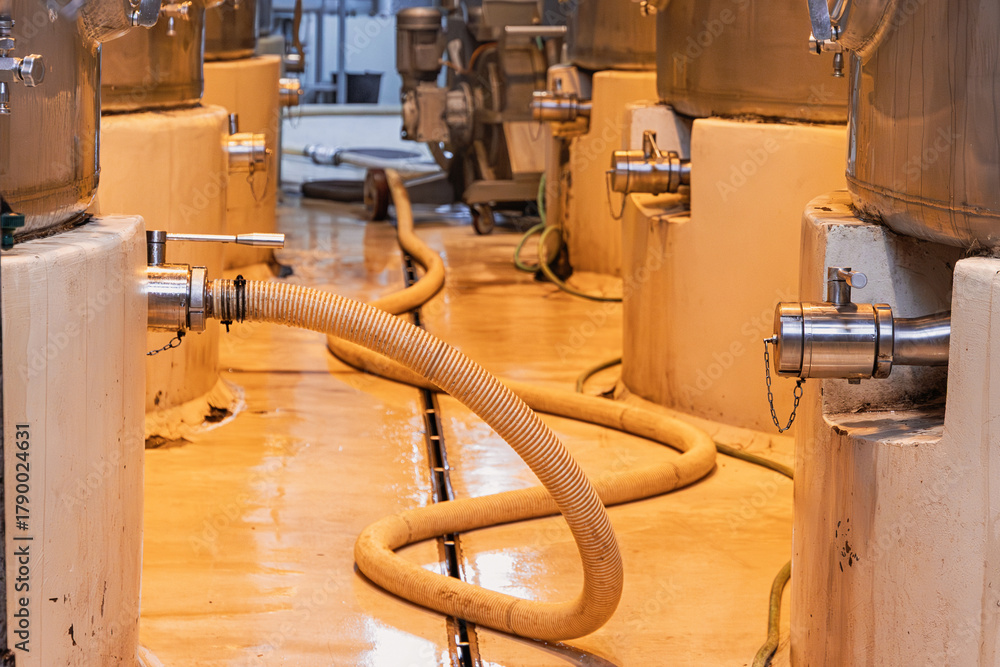 Obraz premium Industrial plant floor showing a row of large processing tanks with stainless steel valves and connected hose, factory environment