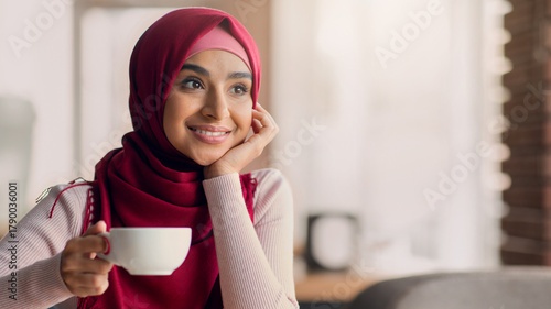 Fotografía A young woman wearing a red hijab sits at a cafe, holding a white cup of coffee