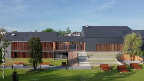 Aerial lateral movement along modern slate and timber-clad public pool, Eisenberga Street Krakow, with sun lounges in landscaped park setting.