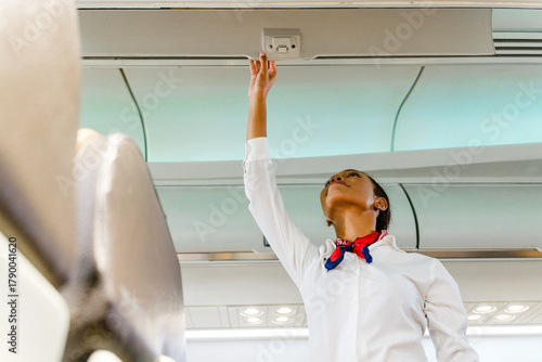 Flight attendant in uniform reaches to adjust an overhead panel inside a modern airplane cabin during preflight checks. Inflight cabin airline service business concept