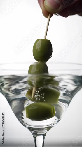 Three vibrant emerald green olives on a polished toothpick gently lowered into a martini glass with gin-clear liquid, creating an ethereal splash against a clean white background. Luxurious product