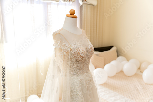 A beautiful wedding dress is showcased on a mannequin in a bright, inviting room. Soft light filters through the window, creating a dreamy atmosphere with white balloons adorning the space.