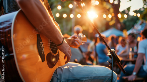 A solo musician playing an acoustic guitar on an outdoor festival stage with a relaxed, intimate crowd drinking beer.