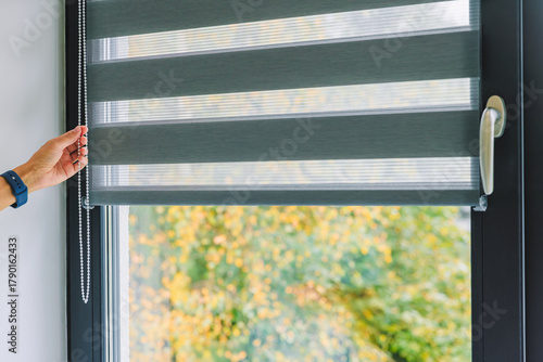 A woman opens black roller blinds on a window in a room. A woman's hand pulls a cord, closing the roller blinds in the room.