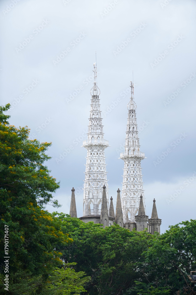 Obraz premium Intricate white Gothic spires of the Jakarta Cathedral, Indonesia, rising above a foreground of lush green trees on a cloudy day
