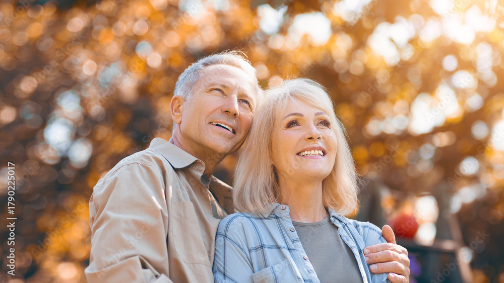 Fototapeta premium A joyful couple stands close together in a park, surrounded by colorful autumn leaves. They share a warm smile as sunlight filters through the trees, creating a serene atmosphere.