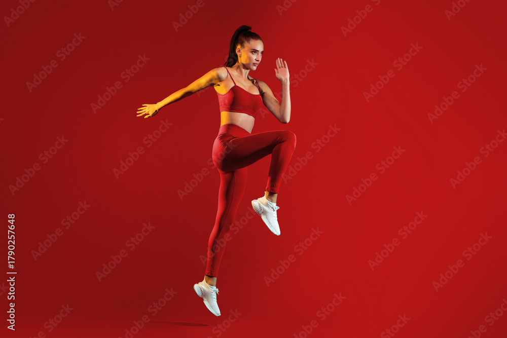 Naklejka premium A fit woman is captured mid-jump in striking red activewear on a vivid red background, showcasing her dynamic movement and athleticism. The scene conveys energy and determination.