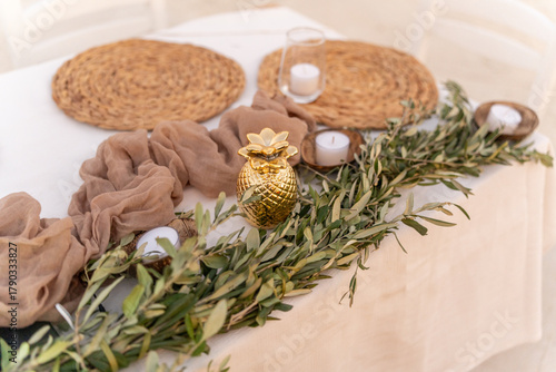 A beautifully arranged table features a golden pineapple centerpiece surrounded by olive branches and lit candles. The setting is inviting for a daytime event, enhancing the atmosphere.