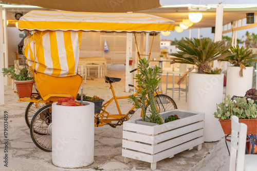 A vibrant yellow bicycle cart sits under a striped canopy, surrounded by potted plants. The outdoor cafe is lively and bright, perfect for enjoying a sunny day by the beach.