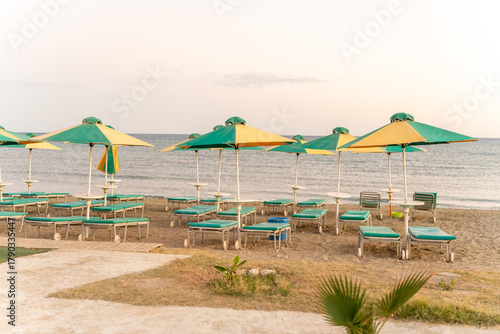 Beach loungers with green cushions sit beneath colorful umbrellas at sunset, offering a peaceful retreat by the shore and inviting relaxation by the ocean.