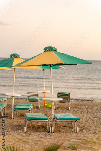 Loungers and umbrellas in green and yellow sit on the sandy beach near the ocean at sunset. The calm water reflects warm colors, creating a serene atmosphere for relaxation.