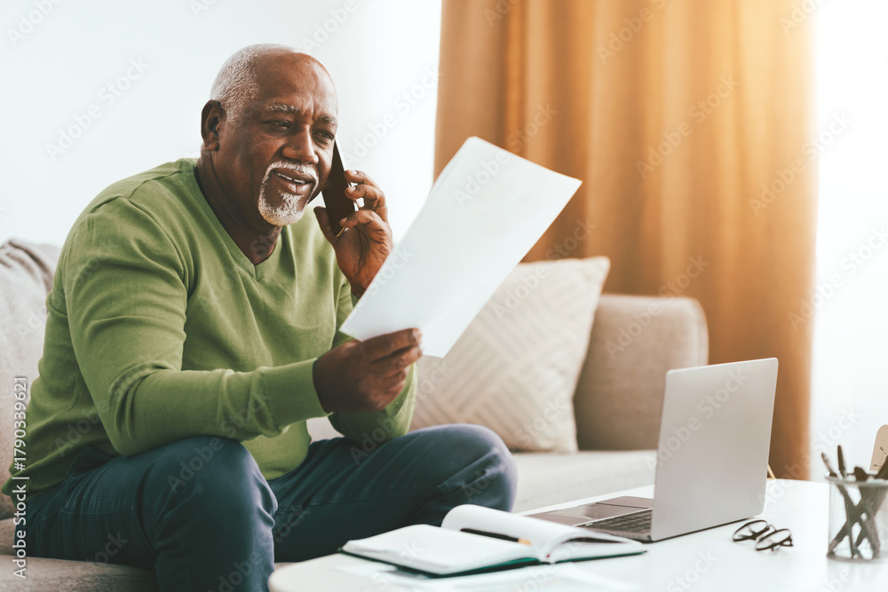 Fototapeta premium A senior man sits comfortably on a couch, holding papers in one hand and talking on the phone. Sunlight streams through the window, creating a warm atmosphere. A laptop rests nearby.
