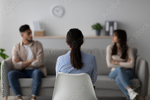 In a therapy session, a female psychologist is assisting a quarreling middle eastern couple. The scene captures the back view of the psychologist as she guides the couple toward resolution.