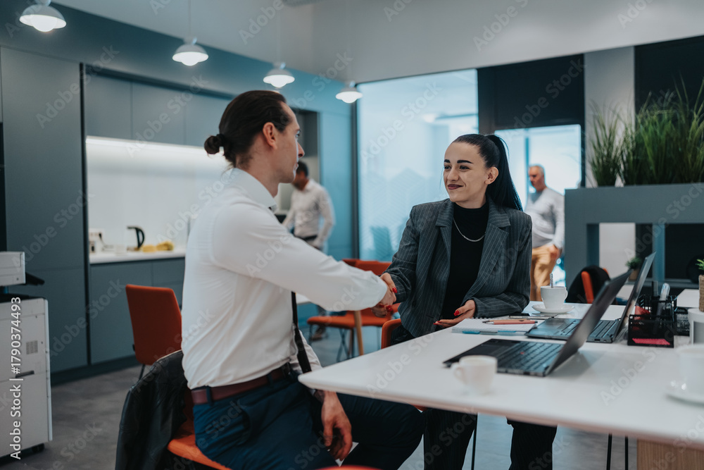 Obraz premium Two professionals greet with a handshake in a bright office setting, surrounded by laptops, coffee, and teamwork.