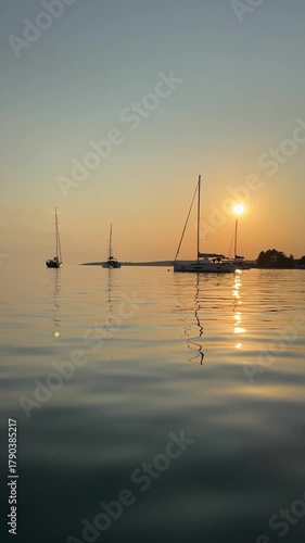 Anchored yachts gently sway on calm water at sunset, viewed from the sea in Croatia. A peaceful Adriatic seascape with glowing silhouettes and soft evening light