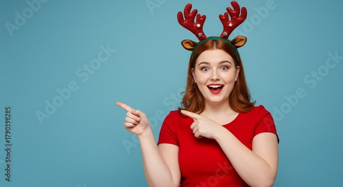 Young woman wearing reindeer antlers and a red shirt, pointing to the side with a surprised expression on a blue background
